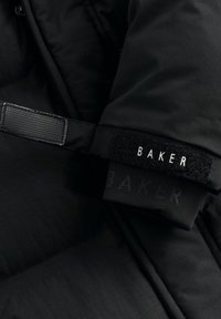 Doudoune noire avec une sangle en velcro affichant le mot "BAKER". Le tissu semble lisse avec une texture douce et un design minimaliste.
