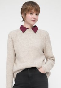 Beige räfflad tröja med rund halsringning, med en vinröd skjorta under. Modellen bär åtsittande svarta jeans.