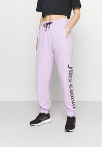 Světle fialové tepláky s elastickým pasem na šňůrku, bočními kapsami a černým текстem podél levé nohy. Na nohou má černé sportovní tenisky.