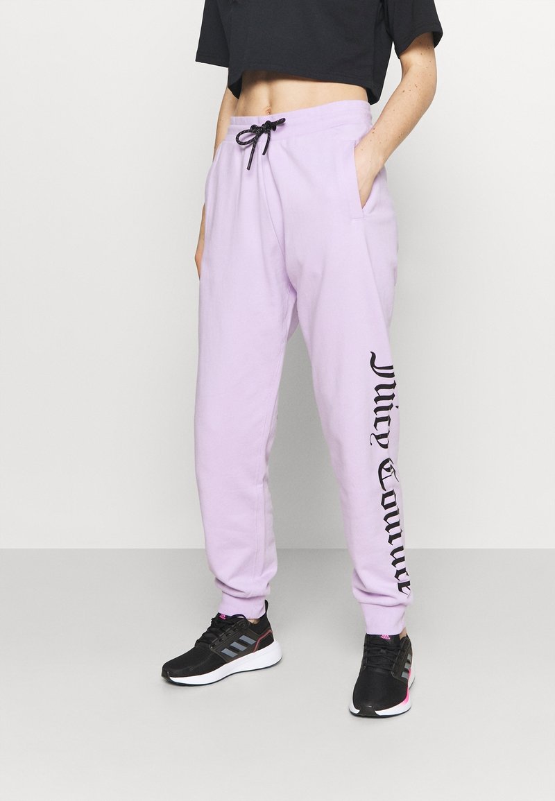 Světle fialové tepláky s elastickým pasem na šňůrku, bočními kapsami a černým текстem podél levé nohy. Na nohou má černé sportovní tenisky.