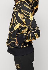 Sweatshirt à capuche de base noire, avec un motif de chaînes dorées et d'étiquettes. Poignets et ourlet côtelés, tissu doux avec une finition brillante.