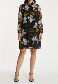 Vestido negro de manga larga con estampado floral colorido, cuello fruncido y sobrecapa transparente. Presenta un corte relajado y llega hasta la mitad del muslo.