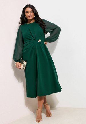 Frau in einem grünen Midikleid mit transparenten langen Ärmeln, goldenen Creolen und einer goldenen Clutch in der Hand posierend vor einer weißen Wand.