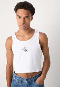 Bijela rebrasta majica bez rukava s krojenim dizajnom, na prednjoj strani s višečlanim "Calvin Klein Jeans" logotipom, u kombinaciji s plavim trappericama.