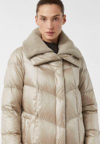 Pufferjack in een lichtbeige kleur met een gewatteerd ontwerp, voorzien van een grote textuur gebreide kraag en een sluiting met knopen aan de voorkant.