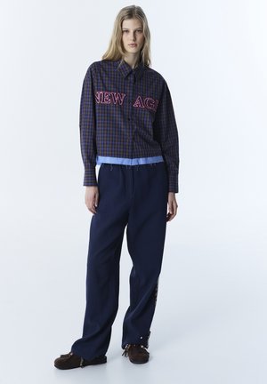 Junge Frau mit blondem Haar trägt ein blau kariertes Hemd mit der Aufschrift "NEW AGE", lockere marineblaue Hosen und braune Mokassin-Schuhe mit Perlen.