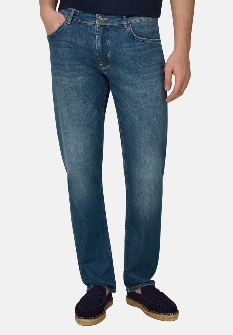 Man draagt slanke blauwe jeans met voorzakken, een knoop- en ritssluiting, gecombineerd met donkere instapschoenen met geweven zolen.