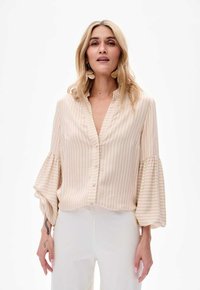 Jasno beżowa bluzka z pionowymi paskami, z przodu na guziki i szerokimi rękawami balonowymi. Gładka faktura, bez kołnierzyka, widoczne białe spodnie.