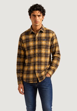 Uomo con camicia a quadri marrone e gialla con taschino sul petto e jeans blu, in piedi con una mano in tasca.