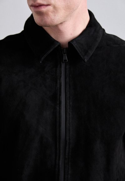 Veste en daim noire avec un col, disposant d'une fermeture éclair à l'avant et d'une surface texturée. Gros plan sur la zone de la poitrine.