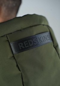 Veste rembourrée vert olive avec une étiquette noire texturée portant des lettres en relief. Le tissu semble lisse avec une finition mate.