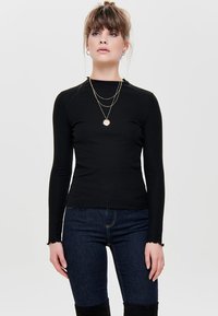 Zwarte geribbelde lange mouwtop met een hoge hals, een getailleerde pasvorm en een iets onafgewerkt zoom, gecombineerd met donkere jeans en gelaagde kettingen.
