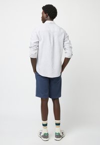 Camisa de manga larga a rayas en azul claro y blanco, pantalones cortos azul marino, con zapatillas deportivas y calcetines de caña corta a rayas. Vista posterior; detalle del cuello visible.