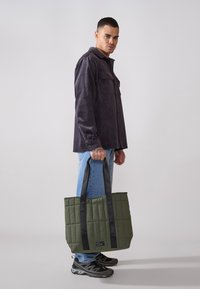 Grønn quiltet tote bag med svarte stropper, med en teksturert overflate og rektangulær form. Svart etikettdetalj nederst.