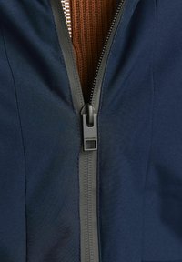 Veste navy avec une fermeture éclair à deux sens et une tirette noire, des accents gris et une doublure en tricot marron texturé visible au niveau du col.