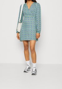 Lyhythihainen mekko sinisissä ja vihreissä geometrisissä kuvioissa, jossa on V-pääntie ja kapeampi vyötärö. Yhdistetty harmaisiin tennareihin ja valkoisiin sukkiin.