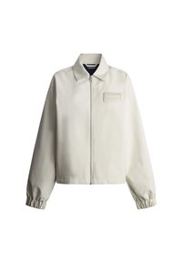 Beige jas met rits, elastische manchetten, puntige kraag en een klein rechthoekig patchlogo op de linkerborst.