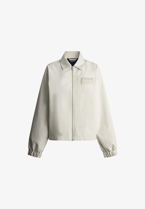 Béžová bunda na zips s elastickými manžetami, špicatým golierom a malým obdĺžnikovým logo nášivkou na ľavej strane hrudníka.