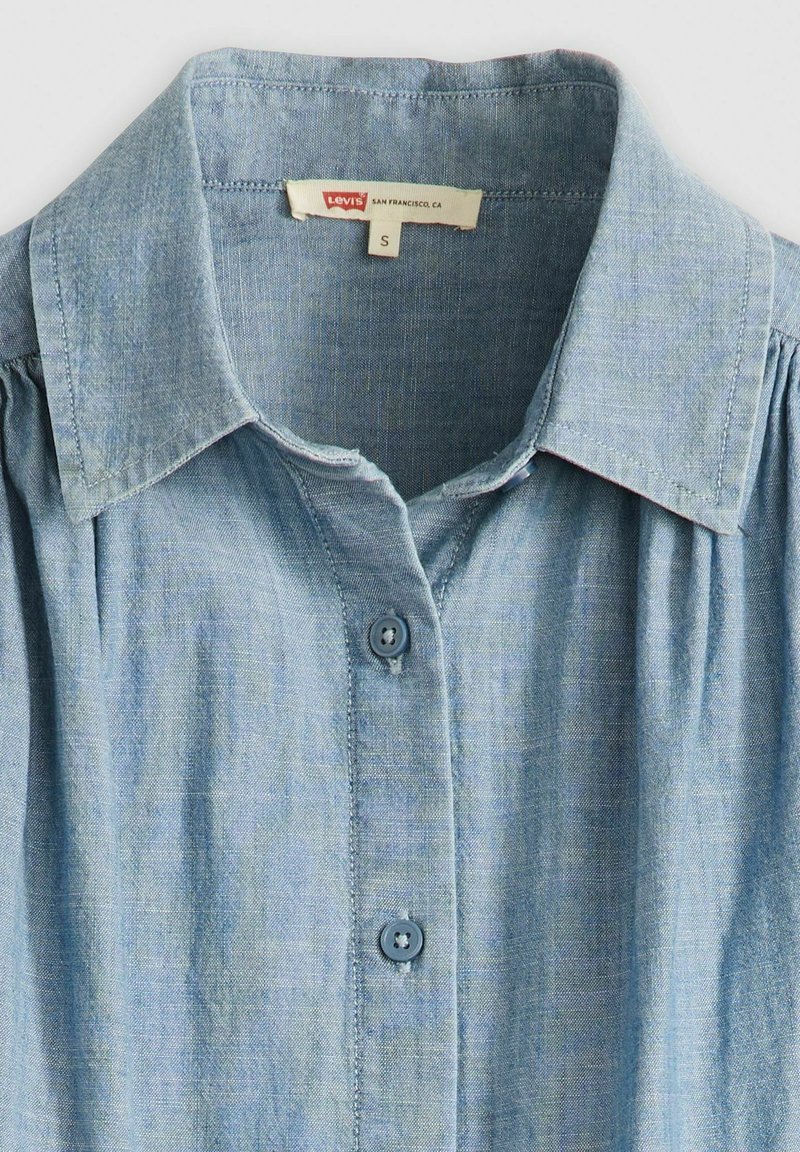 Chemise en denim bleu clair avec un col boutonné, présentant de subtils plis verticaux, une texture de tissu, et une étiquette de taille marquée 'S'.