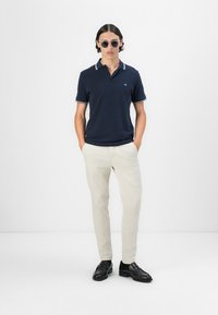 Mornarska polo majica s bijelim detaljima na ovratniku, uparena sa svijetlosmešanim hlačama. Crne cipele upotpunjuju izgled. Model stoji pred bijelom pozadinom.