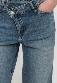 Ανοιχτό μπλε τζιν με ίσια γραμμή, διαθέτει μπροστινή τσέπη και ασημένιο κουμπί. Υφασμάτινη υφή με ορατή ραφή.