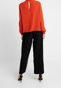 Blouse rouge à manches longues avec poignets froncés, détail goutte d'eau dans le dos et coupe décontractée. Portée avec un pantalon noir à jambes larges et des talons noirs.