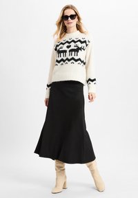 Strickpullover in Creme mit schwarzen Mustern von Rentieren und Zickzack; kombiniert mit einem schwarzen ausgestellten Rock und beigen kniehohen Stiefeln.