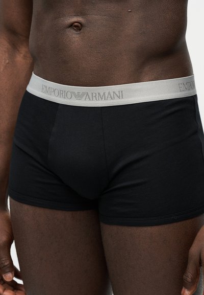 Meessoost torso, kes kannab musti bokserpükse, millel on hõbedane Emporio Armani vöörihm, seisab ühevärvilise valge tausta ees.