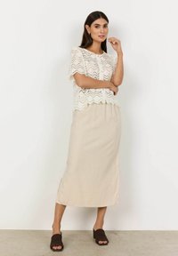 Top en dentelle blanche avec un motif en zigzag, associé à une jupe beige longueur genou. Le modèle porte des sandales à enfiler marron. Design simple et texturé.