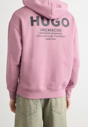Person iført en pink hættetrøje med "HUGO" og tekstdetaljer trykt på ryggen, kombineret med lyseblå denim jeans.
