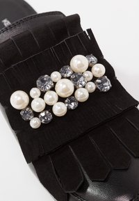 Svart mockaskläm sandal med en fransdesign prydd med en klunga av vita pärlor i olika storlekar och grå rhinestones.