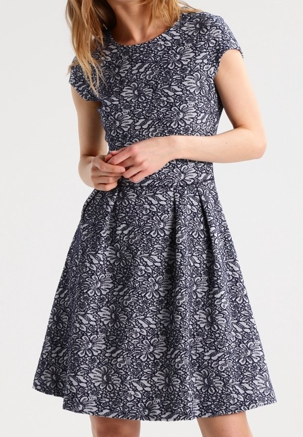 Femme portant une robe courte aux manches courtes et longueur au genou, avec un motif en dentelle florale bleu marine et blanc, debout les mains jointes devant elle.