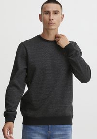 Schwarzer Sweatshirt mit Textur, Rundhalsausschnitt und langen Ärmeln, mit gerippten Bündchen und Saum, getragen mit blauen Jeans. Subtiles Logo-Detail vorhanden.