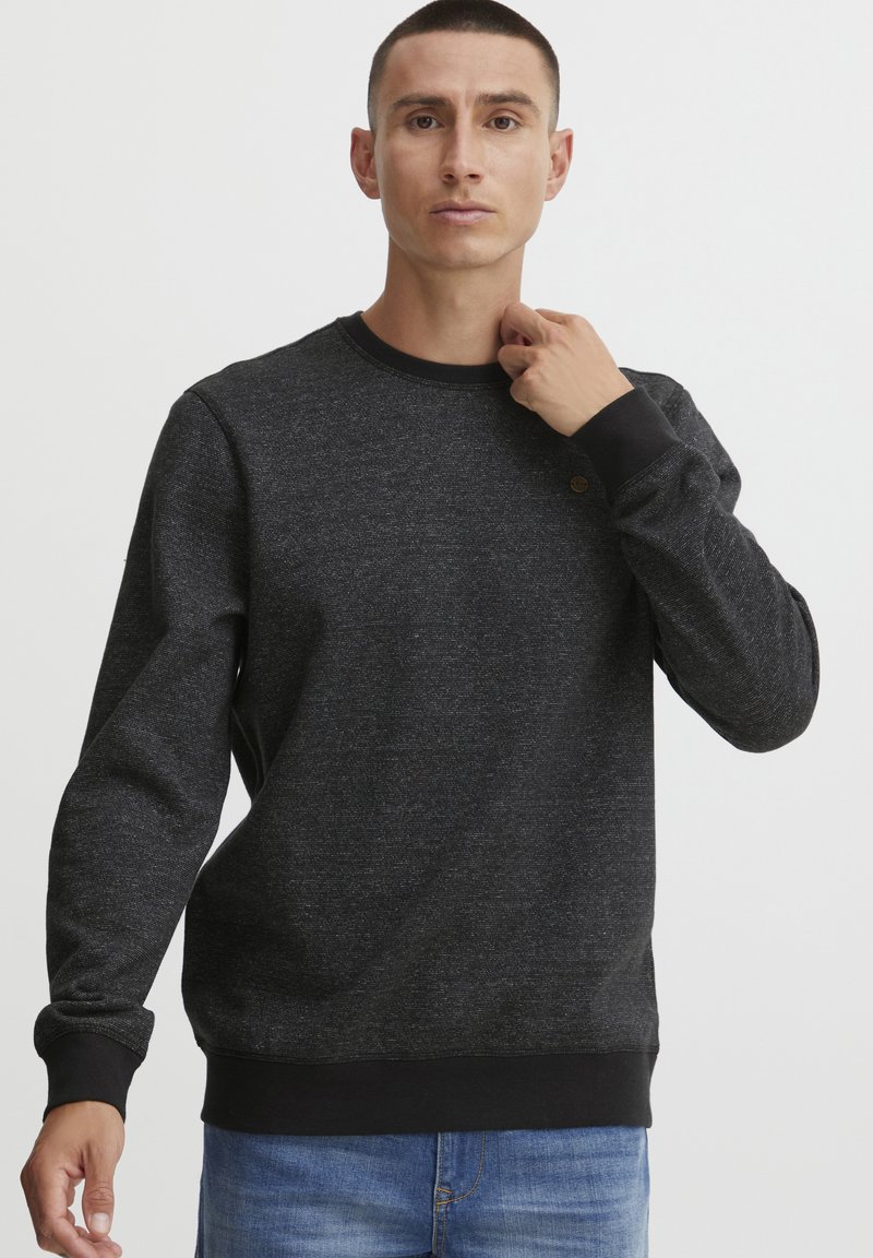 Schwarzer Sweatshirt mit Textur, Rundhalsausschnitt und langen Ärmeln, mit gerippten Bündchen und Saum, getragen mit blauen Jeans. Subtiles Logo-Detail vorhanden.