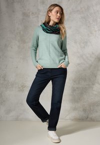 Maglione a maglia verde chiaro con orlo a coste, jeans in denim blu scuro e sneakers bianche. L'outfit include una sciarpa a fantasia attorno al collo.