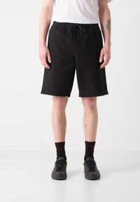 Schwarze Shorts mit Kordelzug und strukturiertem Finish, knielanger Schnitt, Seitentaschen, kombiniert mit schwarzen Sneakern und dunklen Socken.