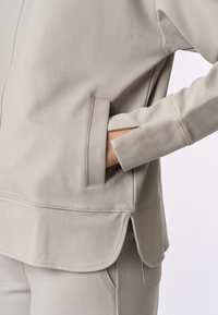 Giacca zip-up grigia chiara con finitura testurizzata, dotata di tasche laterali e orlo curvo. Tessuto morbido, design casual con dettagli di cucitura minimi.