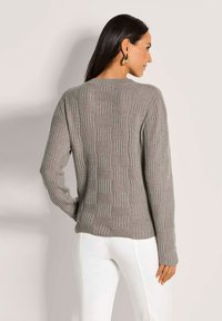 Frau mit langen dunklen Haaren, die einen strukturierten grauen Strickpullover und weiße Hosen trägt, mit dem Rücken zu einem schlichten Hintergrund.