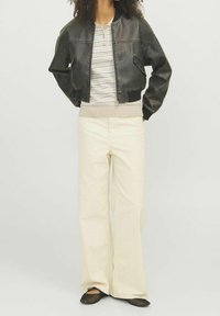 Giubbotto bomber in pelle nera con zip frontale e orlo a costine, polo beige a righe, pantaloni ampi color crema e ballerine in rete nera.