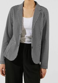 Blazer à motifs noir et blanc avec un col cranté et des manches longues, fabriqué en tissu texturé ; associé à un haut blanc et un bas noir.