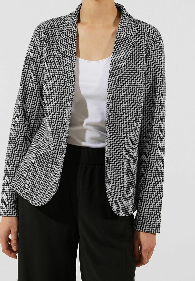Blazer à motifs noir et blanc avec un col cranté et des manches longues, fabriqué en tissu texturé ; associé à un haut blanc et un bas noir.