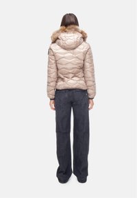Giacca beige imbottita con motivo a rombi, cappuccio foderato in pelliccia e dettagli neri. Indossata sopra pantaloni in denim scuri a gamba larga.