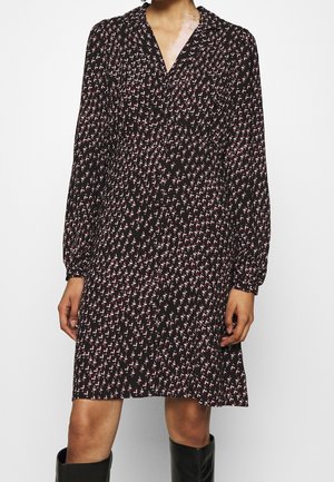 Frau trägt ein schwarzes langärmliges Kleid mit kleinen pinken und weißen Blumenmustern und schwarzen kniehohen Stiefeln.