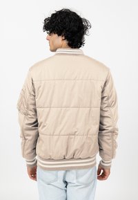 Beżowa kurtka puchowa z prążkowanymi mankietami i dołem, z bocznymi kieszeniami i pikowaną teksturą. Widok z tyłu pokazuje gładki, opływowy design.