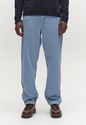 Férfi világoskék egyenes szárú farmernadrágban, sötétbarna bőr cipőben és sötét hosszú ujjú ingben egy egyszerű háttér előtt áll.