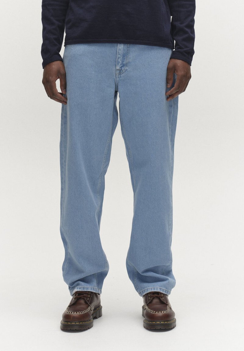 Mann som har på seg lyseblå jeans med rette ben, mørkebrune skinnsko og en mørk langermet skjorte, står foran en ensfarget bakgrunn.