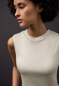 Světlounce šedý tílko bez rukávů s kulatým výstřihem, vyrobené z texturované látky, která jemně třpytí na tmavém pozadí.