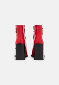 Bottines en vernis rouge avec un talon carré noir, comportant un accent doré sur le côté et une fermeture éclair à l'arrière.