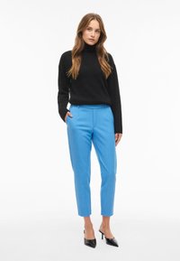 Must-have musternakk, sinised kohandatud püksid, sobiv disain, sile tekstuur, koos mustade kontsakingadega. Modell seisab, üks käsi taskus.