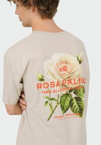 Camisa de algodón beige con un gran gráfico de una rosa blanca, hojas verdes y texto rojo en la parte de atrás. Corte estándar con mangas cortas.